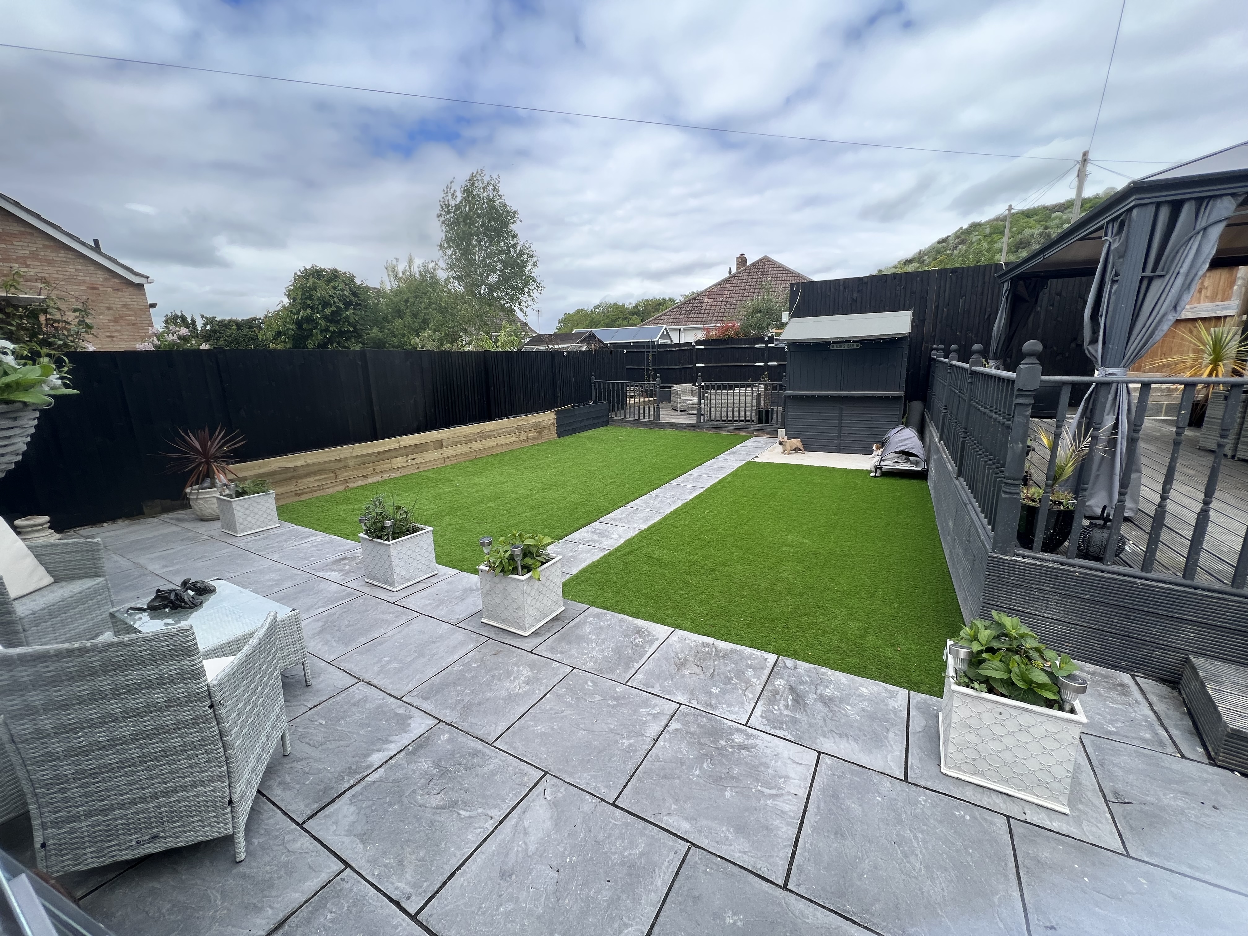 Contemporary Maidstone garden with grey porcelain paving, twin artificial lawns, black fencing and covered decking zone
